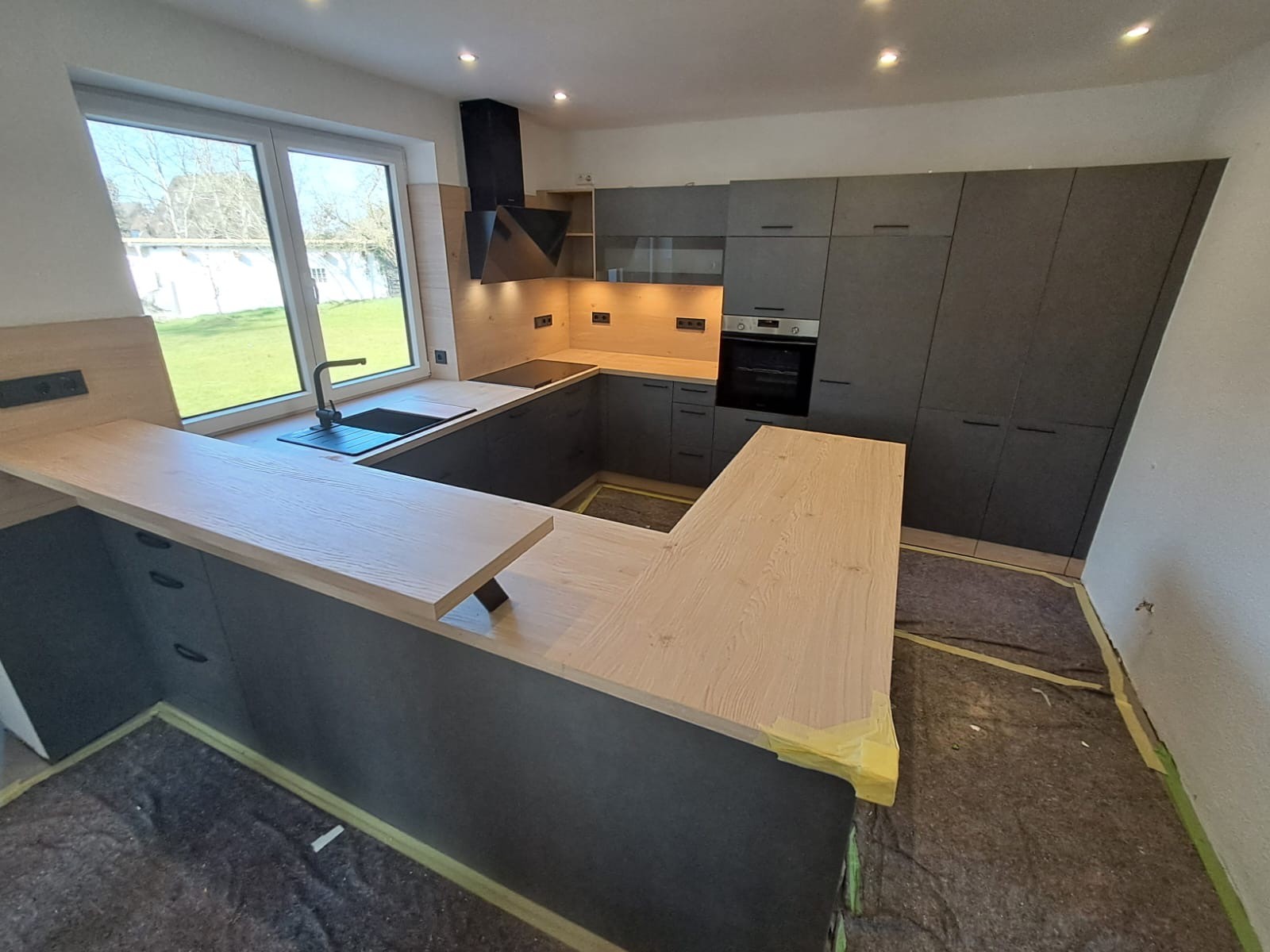 Moderne Küche mit U-förmiger heller Arbeitsplatte und grauen Schränken, großes Fenster.