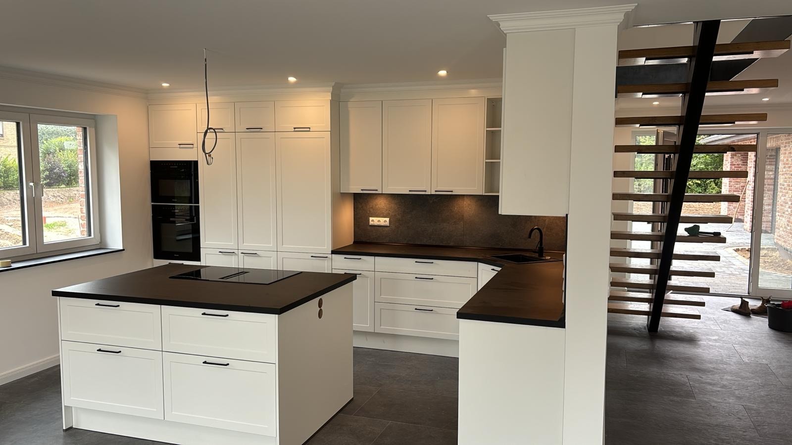 Moderne weiße Küche mit Kochinsel, dunkler Arbeitsplatte und offener Treppe im Hintergrund.