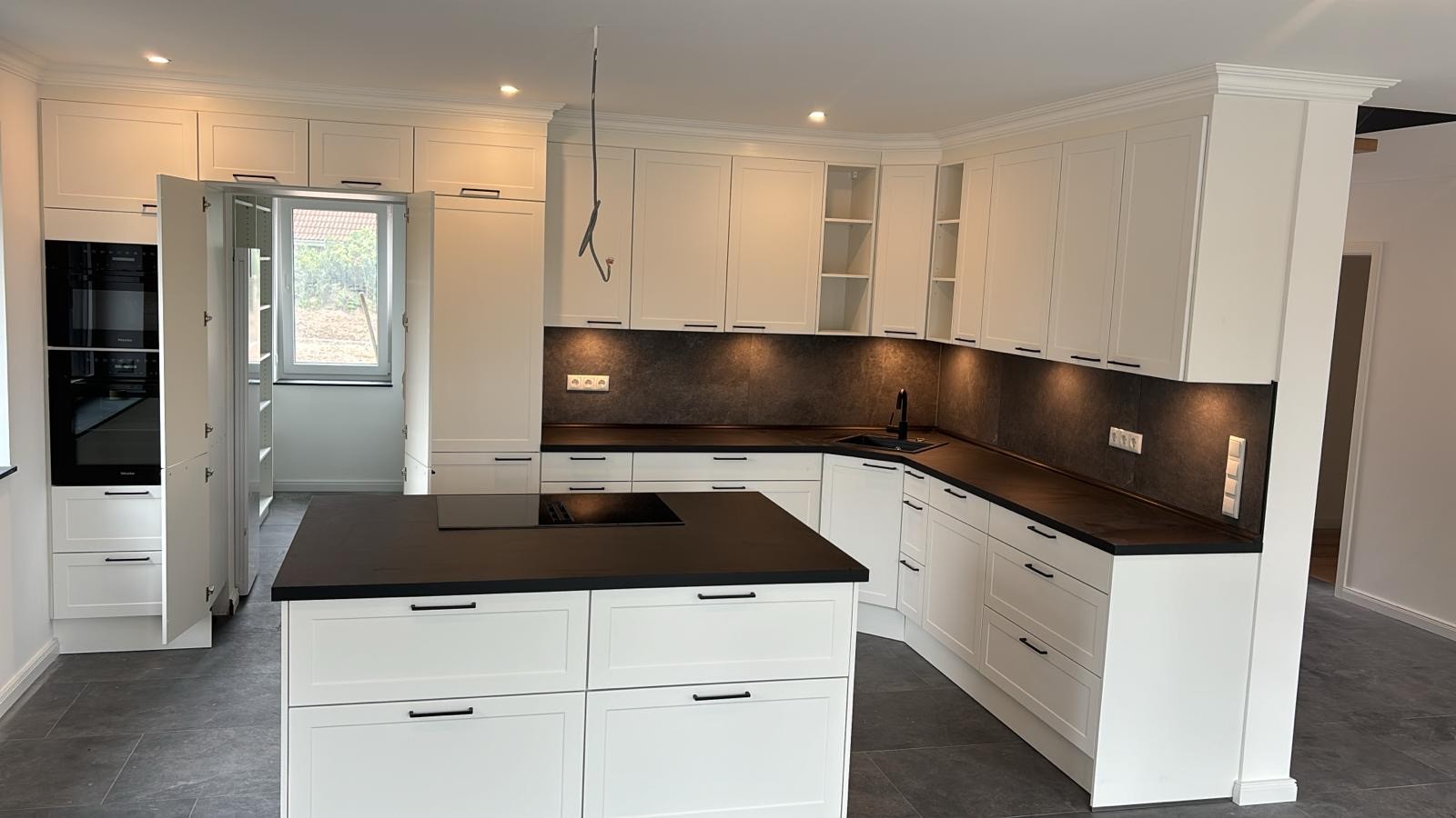 Moderne weiße Küche mit Kochinsel, dunkler Arbeitsplatte und Einbaugeräten.